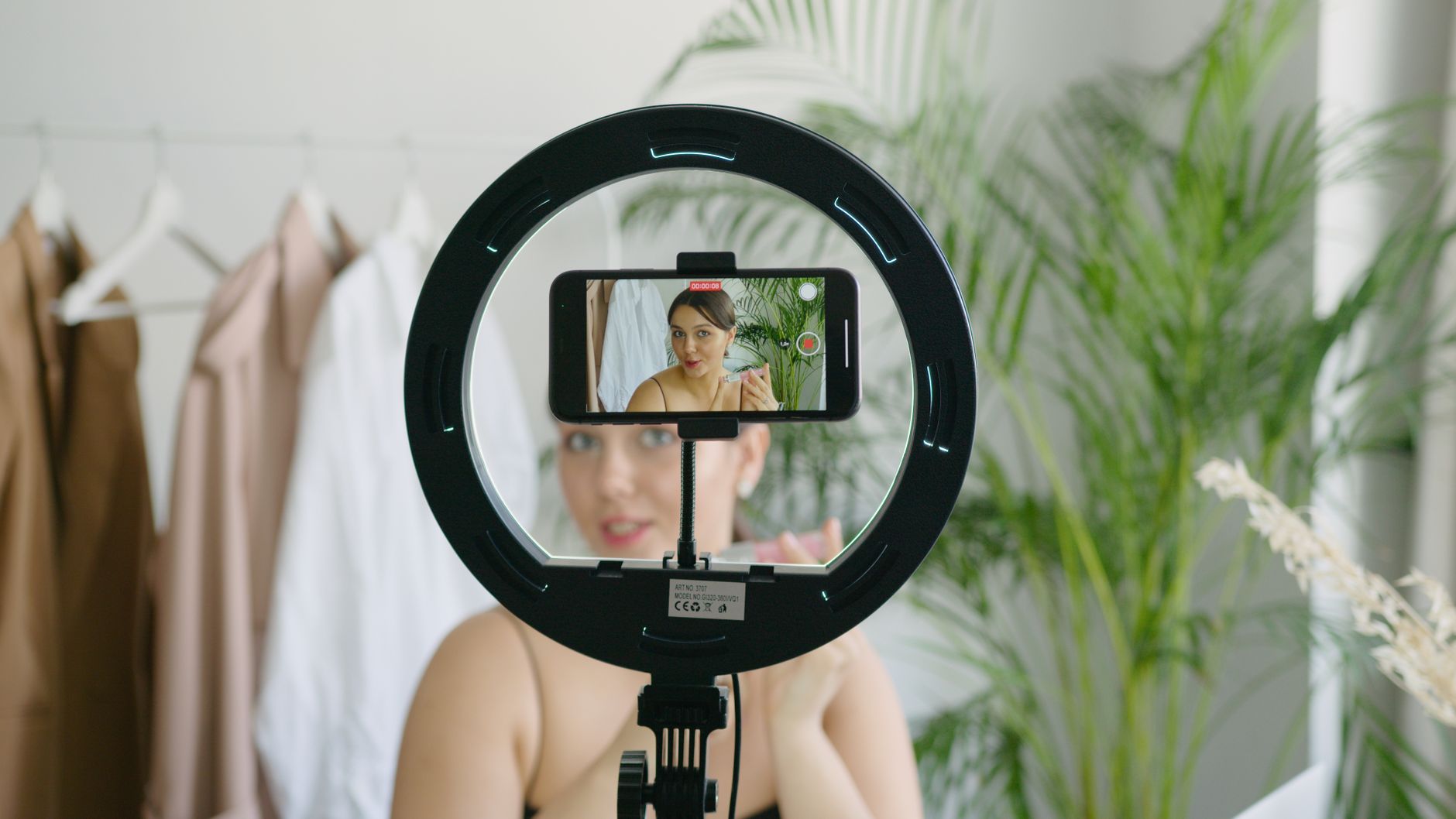 Une femme se filme chez elle avec un smartphone fixé sur un anneau lumineux, devant un portant de vêtements et une grande plante verte